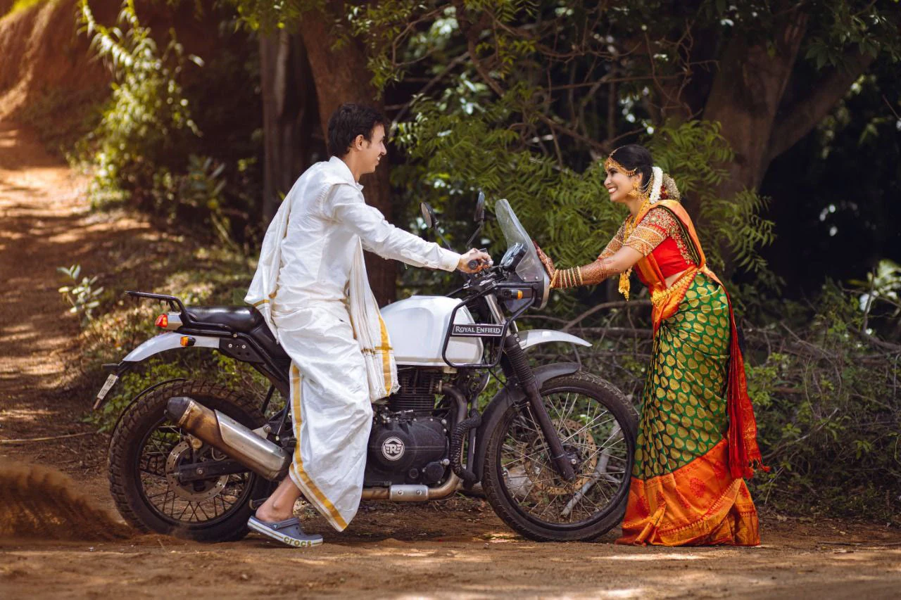 Couple standing on either side of a motorcycle beneath trees