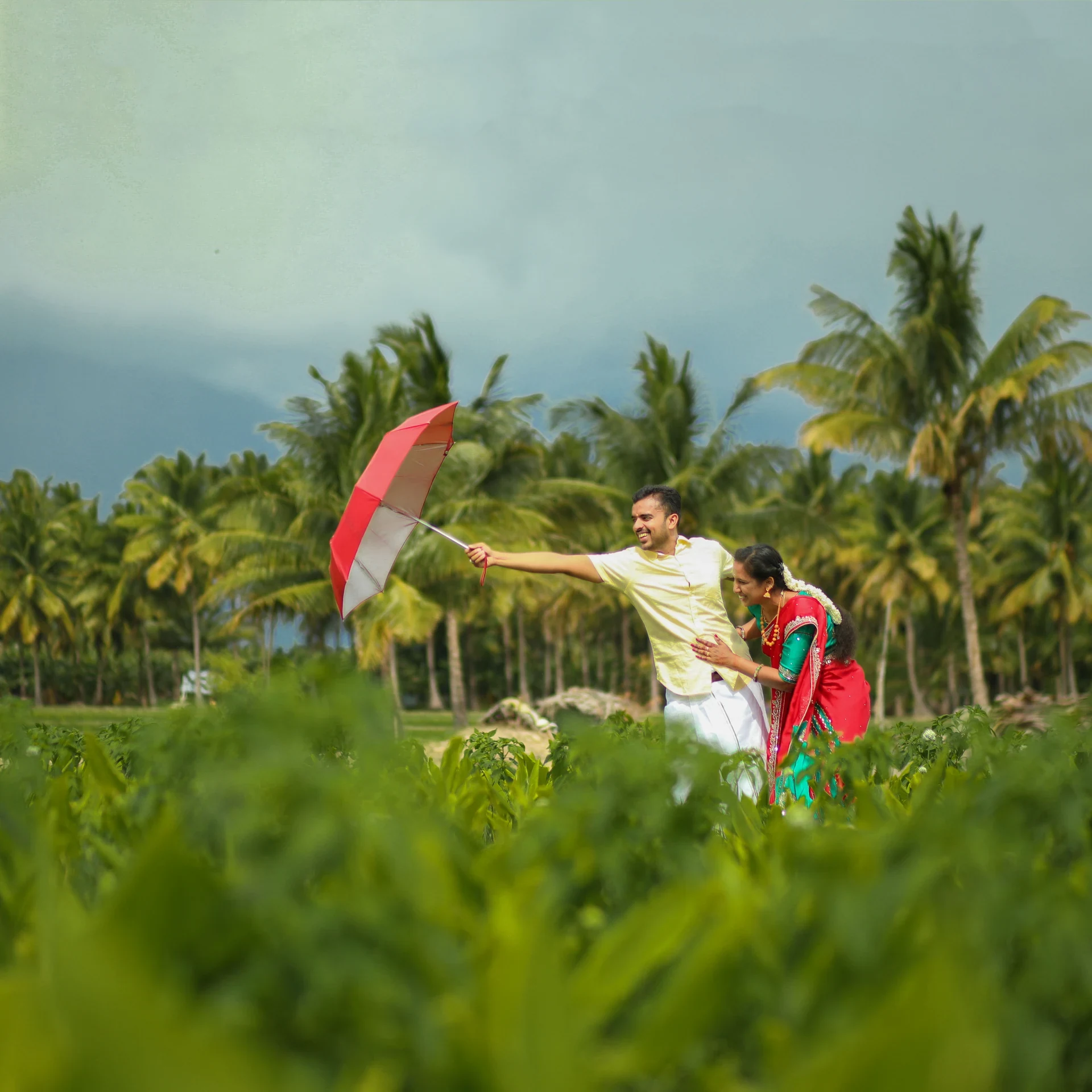 Couple reaching towards a red umbrella against a cloudy sky