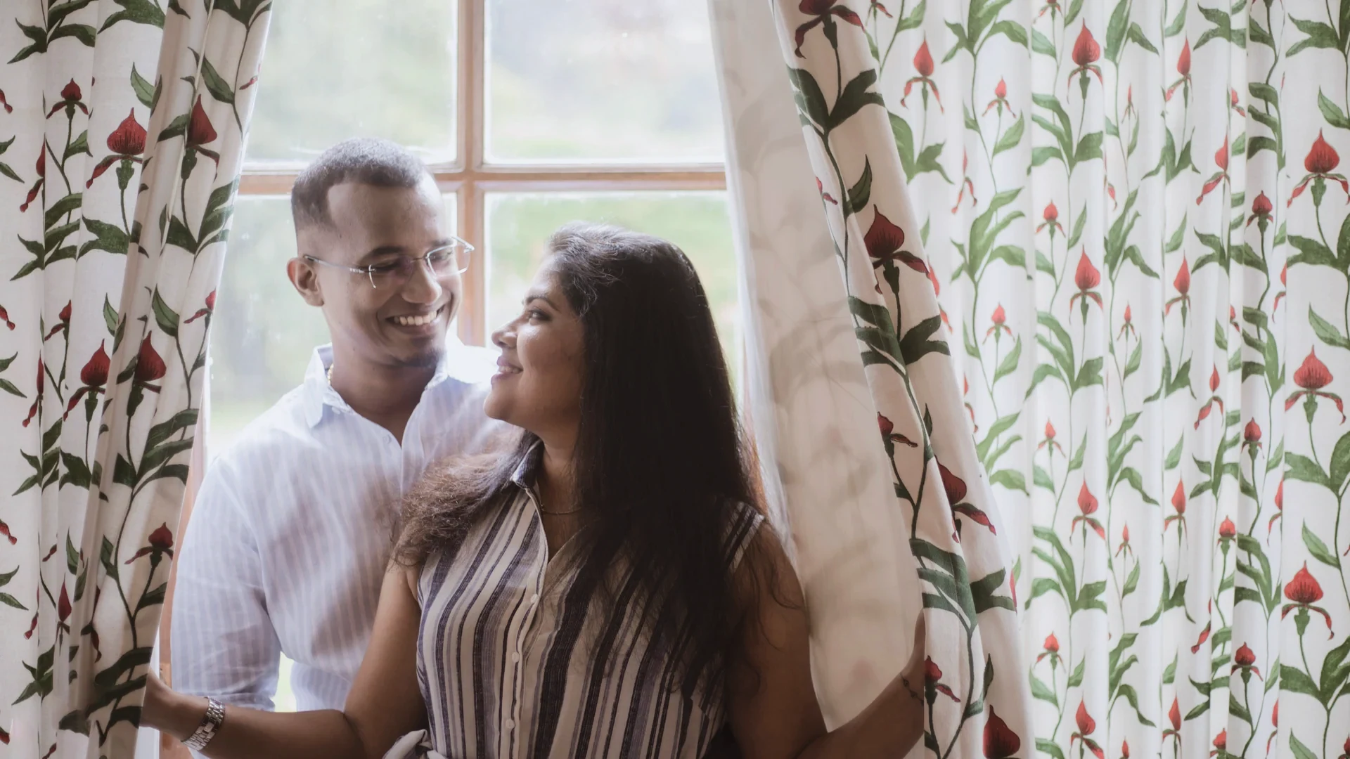 Couple smiling together beside patterned curtains and a bright window